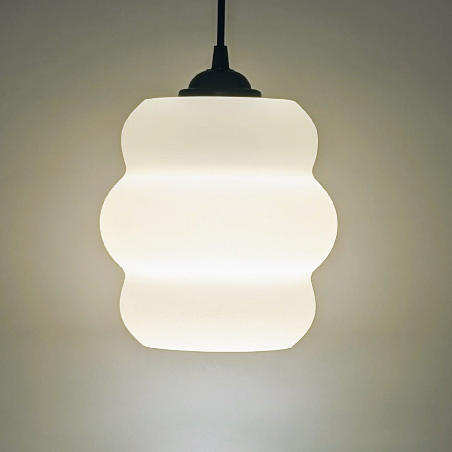 The Mammatus Pendant Light by Dryft Home, crafted from sustainable material, boasts a wavy design that casts a soft white glow. Suspended on a dark cord against a light backdrop, it enhances any space with its minimalist elegance.