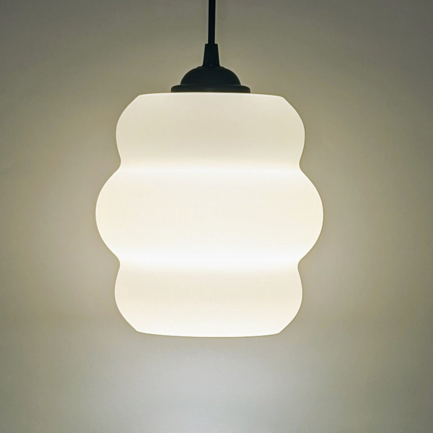 The Mammatus Pendant Light by Dryft Home, crafted from sustainable material, boasts a wavy design that casts a soft white glow. Suspended on a dark cord against a light backdrop, it enhances any space with its minimalist elegance.