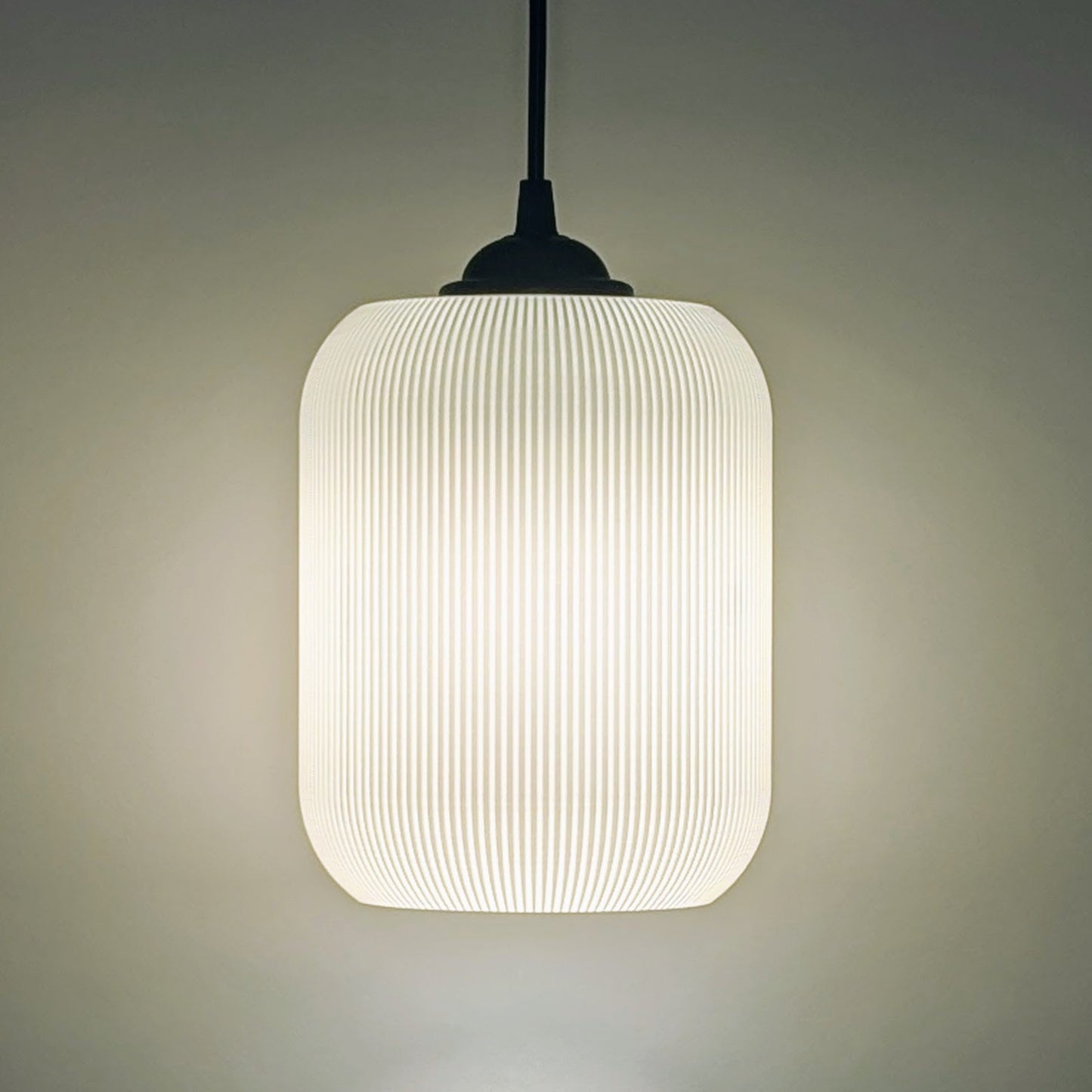 The Cumulis Pendant Light by Dryft Home features a modern cylindrical design with a ribbed translucent shade, crafted using 3D printing. Hanging from a black cord, it emits a soft warm glow against any neutral backdrop.