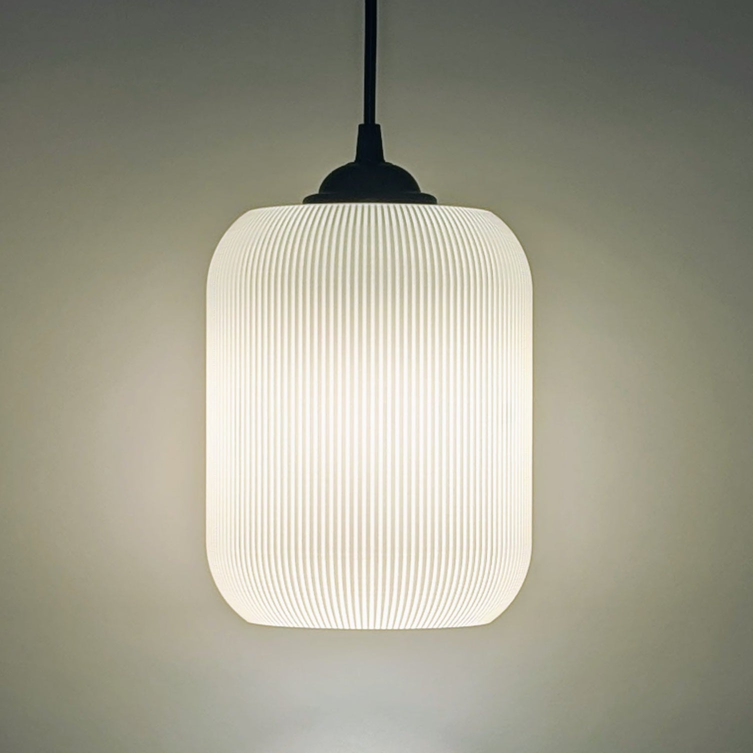The Cumulis Pendant Light by Dryft Home features a modern cylindrical design with a ribbed translucent shade, crafted using 3D printing. Hanging from a black cord, it emits a soft warm glow against any neutral backdrop.