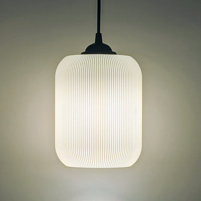The Cumulis Pendant Light by Dryft Home features a modern cylindrical design with a ribbed translucent shade, crafted using 3D printing. Hanging from a black cord, it emits a soft warm glow against any neutral backdrop.