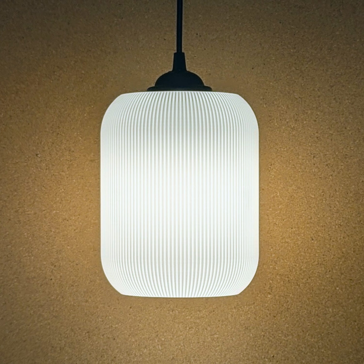 The Cumulis Pendant Light by Dryft Home boasts a ribbed, white cylindrical shade made with 3D printing and sustainable materials. It hangs on a sleek black cord against a beige backdrop, casting a warm glow perfect for modern spaces.