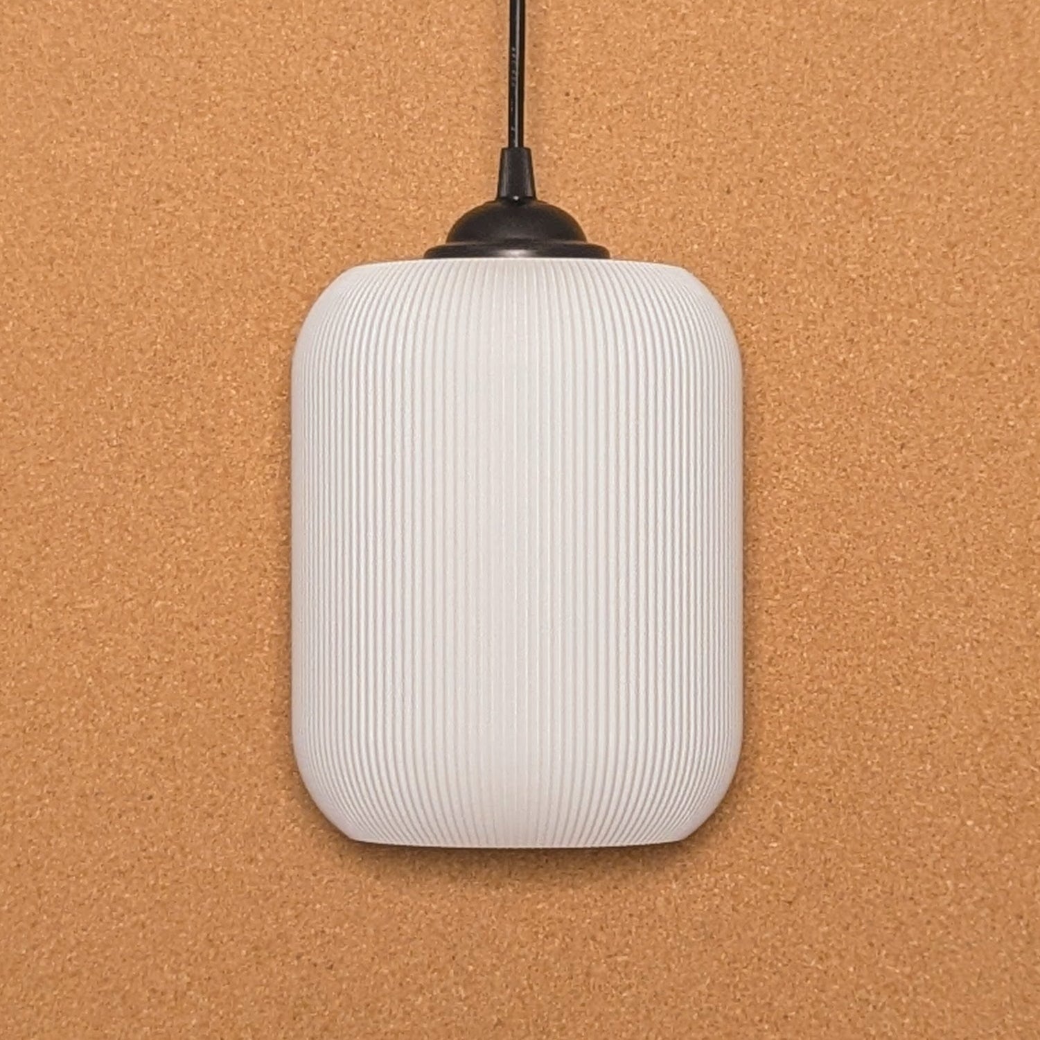 Crafted from sustainable materials using 3D printing, Dryft Homes Cumulis Pendant Light features a ribbed white cylindrical shade on a black cord, set against a textured cork background.