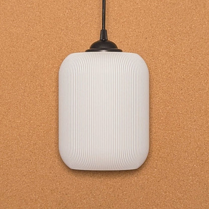 Crafted from sustainable materials using 3D printing, Dryft Homes Cumulis Pendant Light features a ribbed white cylindrical shade on a black cord, set against a textured cork background.