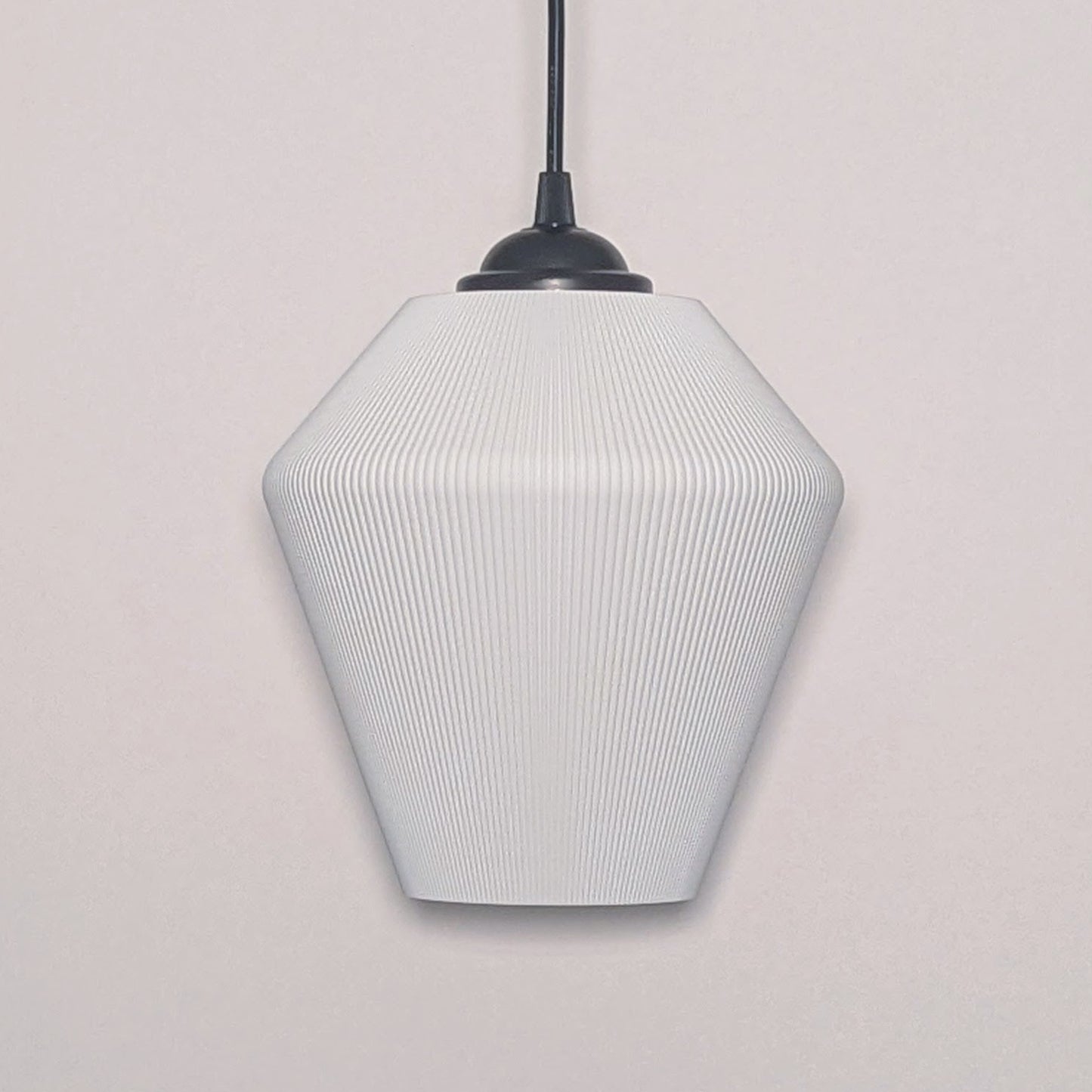 Dryft Homes Inversis Pendant Light showcases a modern geometric white design suspended by a black cord. Crafted sustainably with a 3D printed lampshade, its subtle ribbed texture adds a stylish, minimalist touch to any plain white wall.