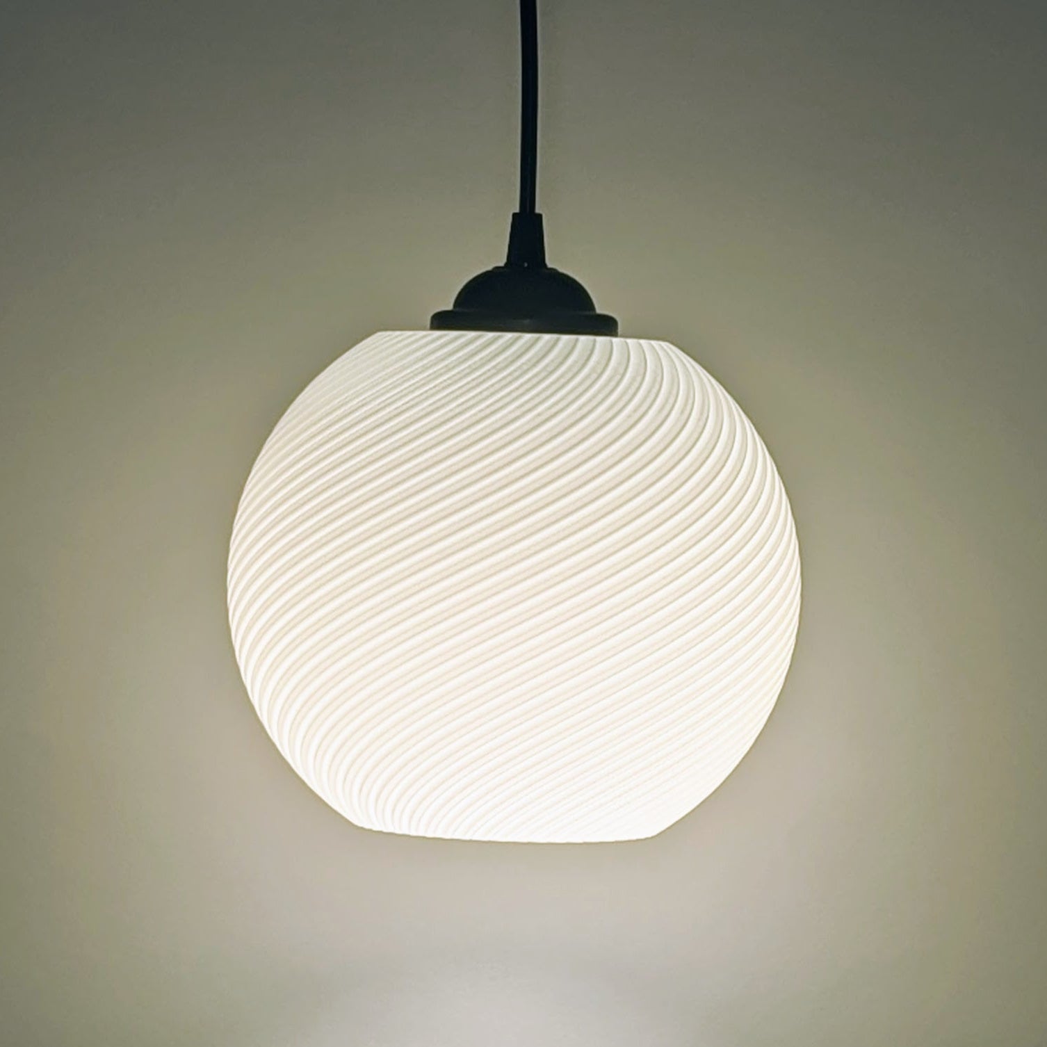 The Vortice Pendant Light by Dryft Home features a 3D printed, swirling pattern. Crafted from biodegradable PLA plastic, it emits a soft glow and hangs from a dark cord, exemplifying modern, sustainable lighting against a neutral background.