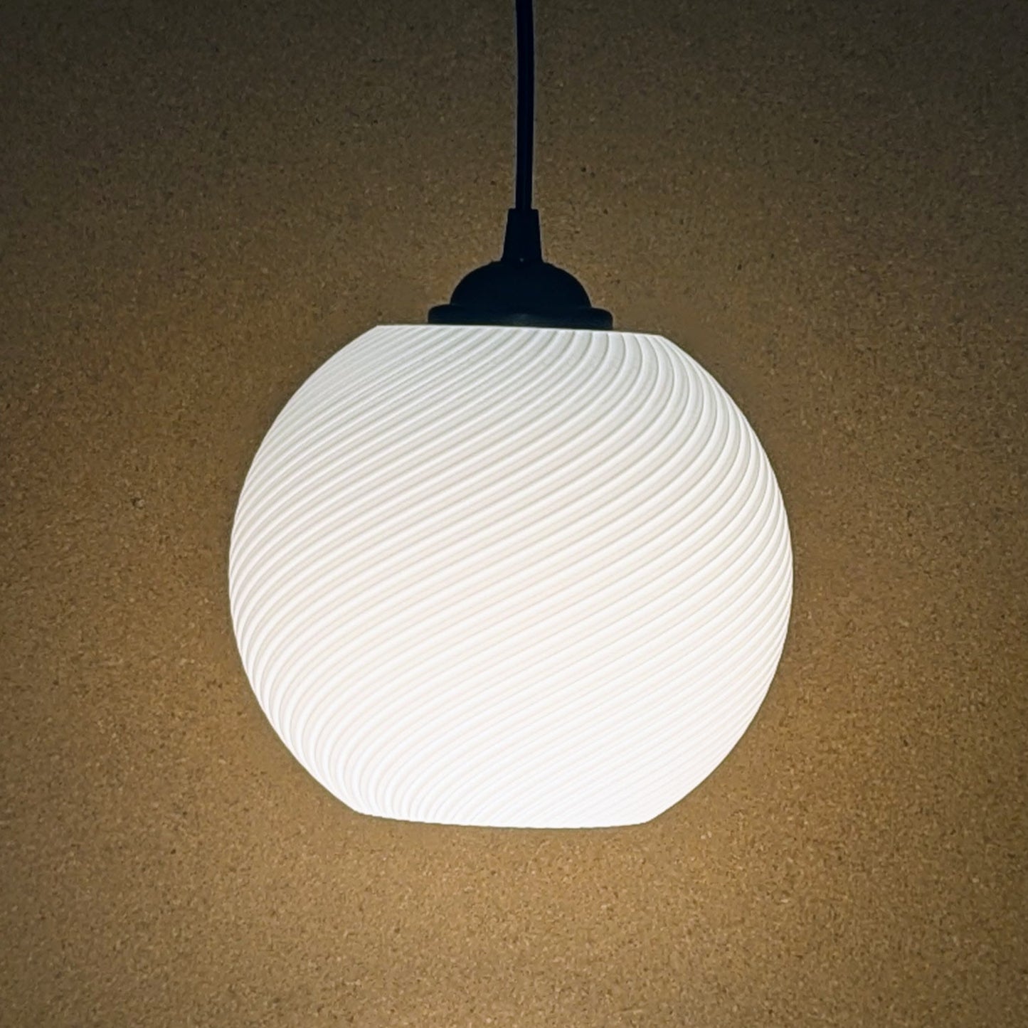 The Vortice Pendant Light by Dryft Home is a 3D printed lamp with a textured, wavy design that casts a soft, warm light on a beige backdrop. Made from biodegradable PLA plastic, it features a black fitting and cord, exemplifying sustainable lighting.