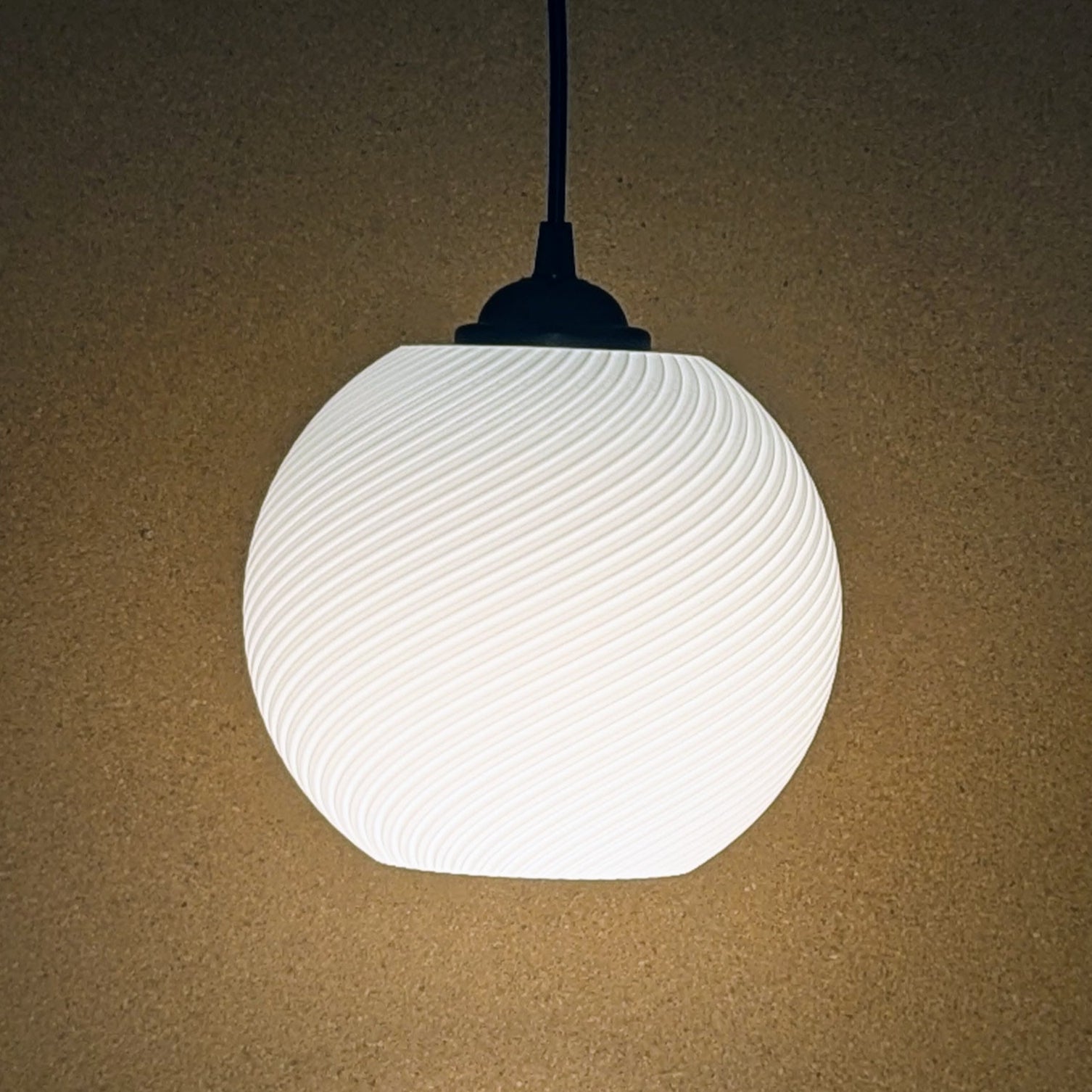 The Vortice Pendant Light by Dryft Home is a 3D printed lamp with a textured, wavy design that casts a soft, warm light on a beige backdrop. Made from biodegradable PLA plastic, it features a black fitting and cord, exemplifying sustainable lighting.