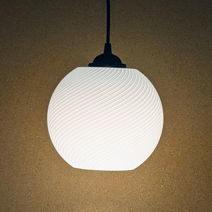 The Vortice Pendant Light by Dryft Home is a 3D printed lamp with a textured, wavy design that casts a soft, warm light on a beige backdrop. Made from biodegradable PLA plastic, it features a black fitting and cord, exemplifying sustainable lighting.