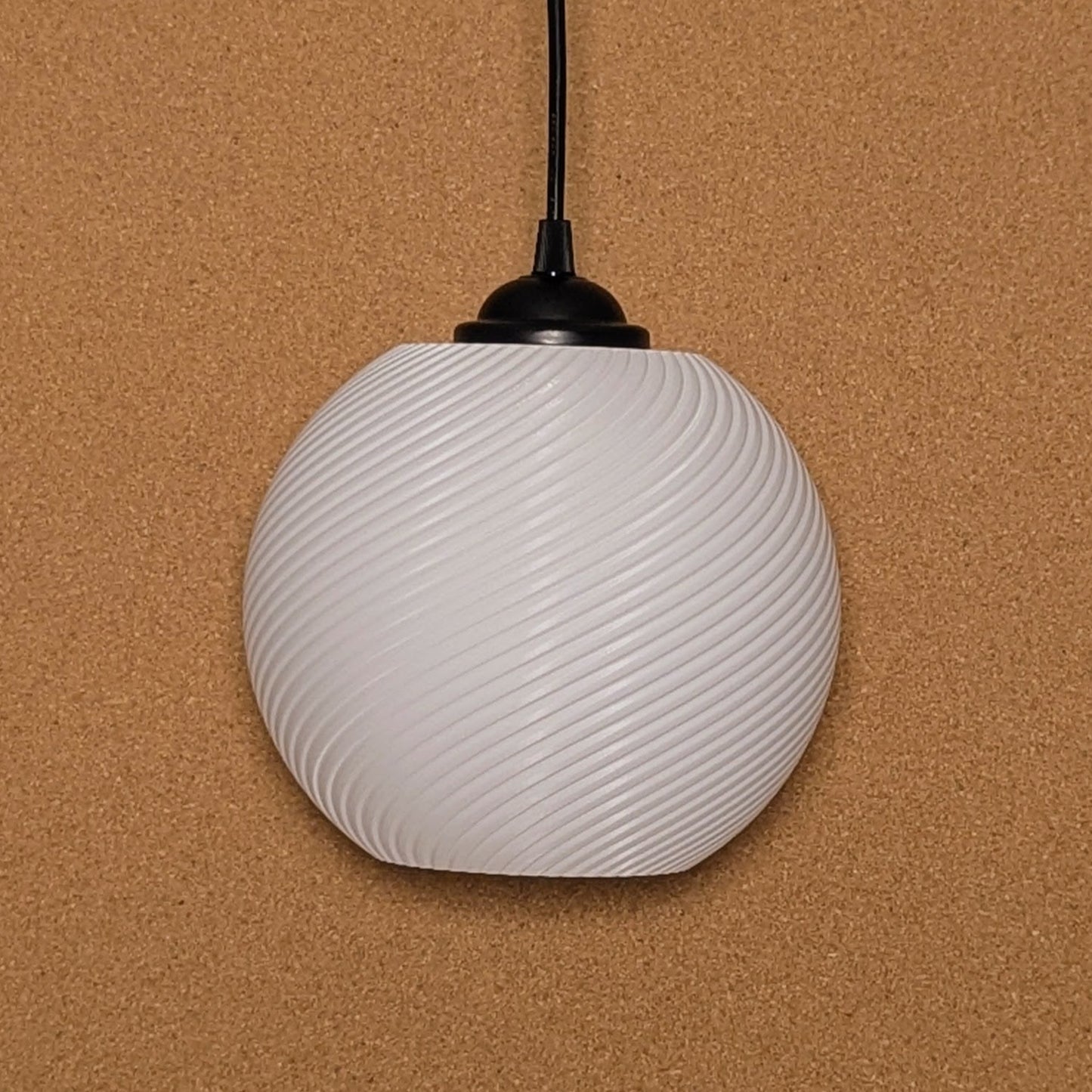 The Vortice Pendant Light by Dryft Home, made of biodegradable PLA plastic, features a spherical, white design with a spiral pattern. It hangs elegantly against a textured brown background with a black top attachment and an extending cord.