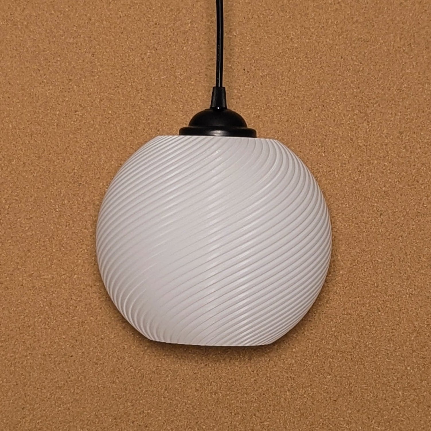 The Vortice Pendant Light by Dryft Home, made of biodegradable PLA plastic, features a spherical, white design with a spiral pattern. It hangs elegantly against a textured brown background with a black top attachment and an extending cord.