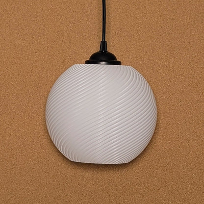The Vortice Pendant Light by Dryft Home, made of biodegradable PLA plastic, features a spherical, white design with a spiral pattern. It hangs elegantly against a textured brown background with a black top attachment and an extending cord.