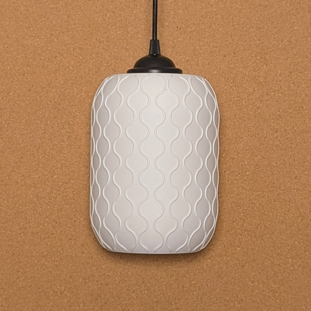 The Cirrus Pendant Light by Dryft Home features a white, textured, cylindrical design with a wavy pattern against a corkboard backdrop. Made from sustainable materials, it hangs elegantly with a black cord, showcasing eco-friendly elegance.