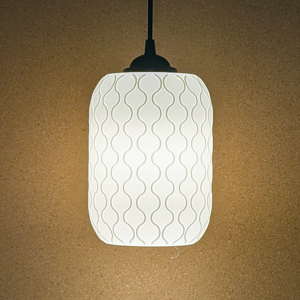 The Cirrus Pendant Light by Dryft Home, featuring a 3D-printed cylindrical white lampshade with a wavy pattern, casts a soft, warm glow against a plain light brown wall. Its crafted from sustainable materials.