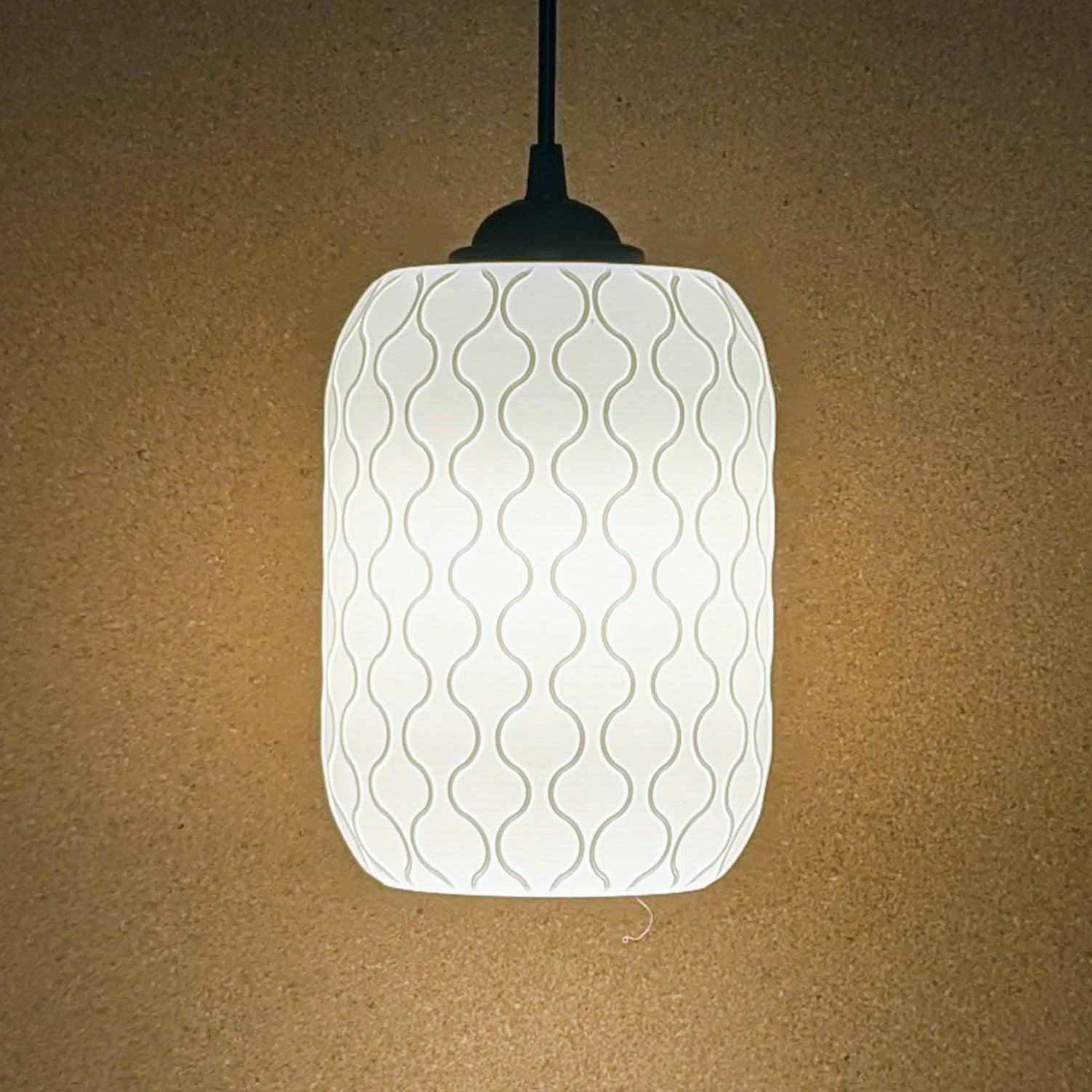 The Cirrus Pendant Light by Dryft Home, featuring a 3D-printed cylindrical white lampshade with a wavy pattern, casts a soft, warm glow against a plain light brown wall. Its crafted from sustainable materials.