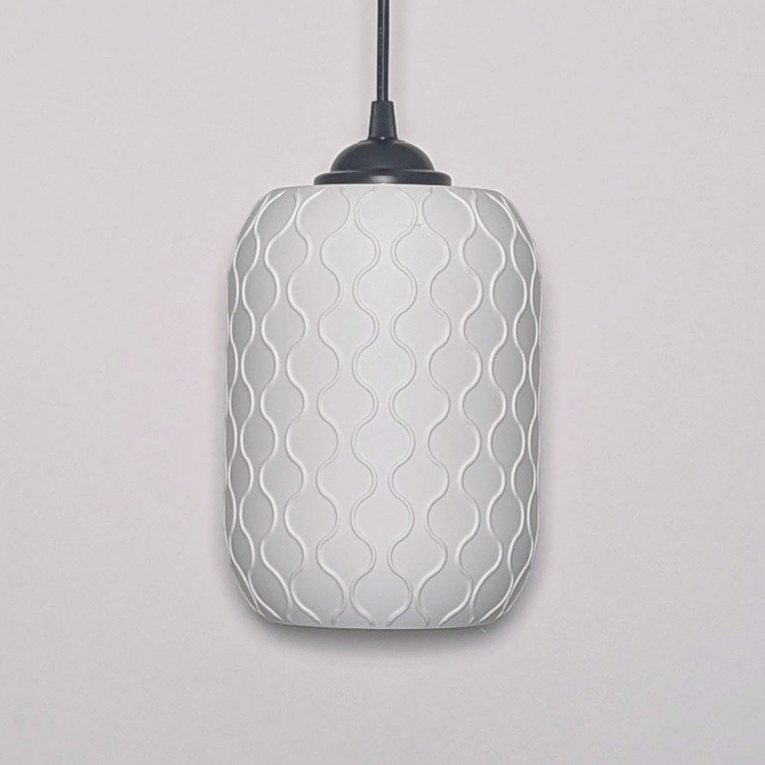 The Cirrus Pendant Light by Dryft Home is crafted from sustainable materials, featuring a smooth, cylindrical white shade. It hangs from a black cord and displays an intricately textured pattern of interlocking wavy lines, creating a subtle geometric design against the wall.