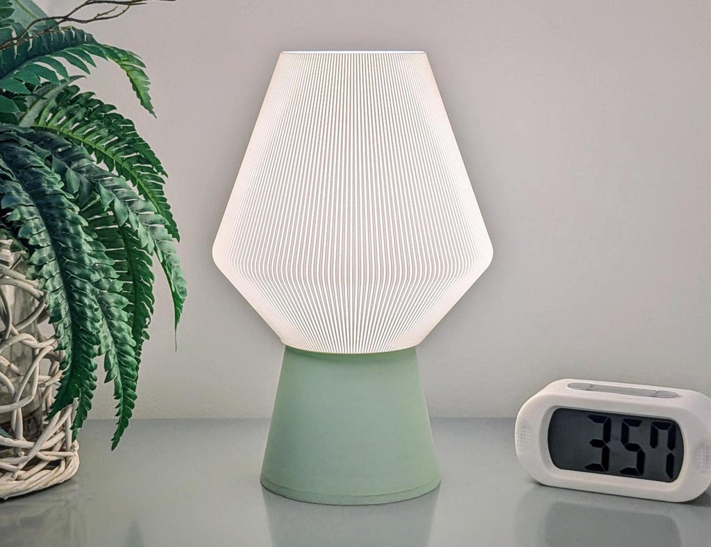 The Inversis Table Lamp by Dryft Home, featuring a pleated white shade and a green biodegradable PLA base, rests on a light surface. A leafy plant in a woven basket sits to the left, and to the right is a digital clock showing 3:59, exemplifying sustainable decor.