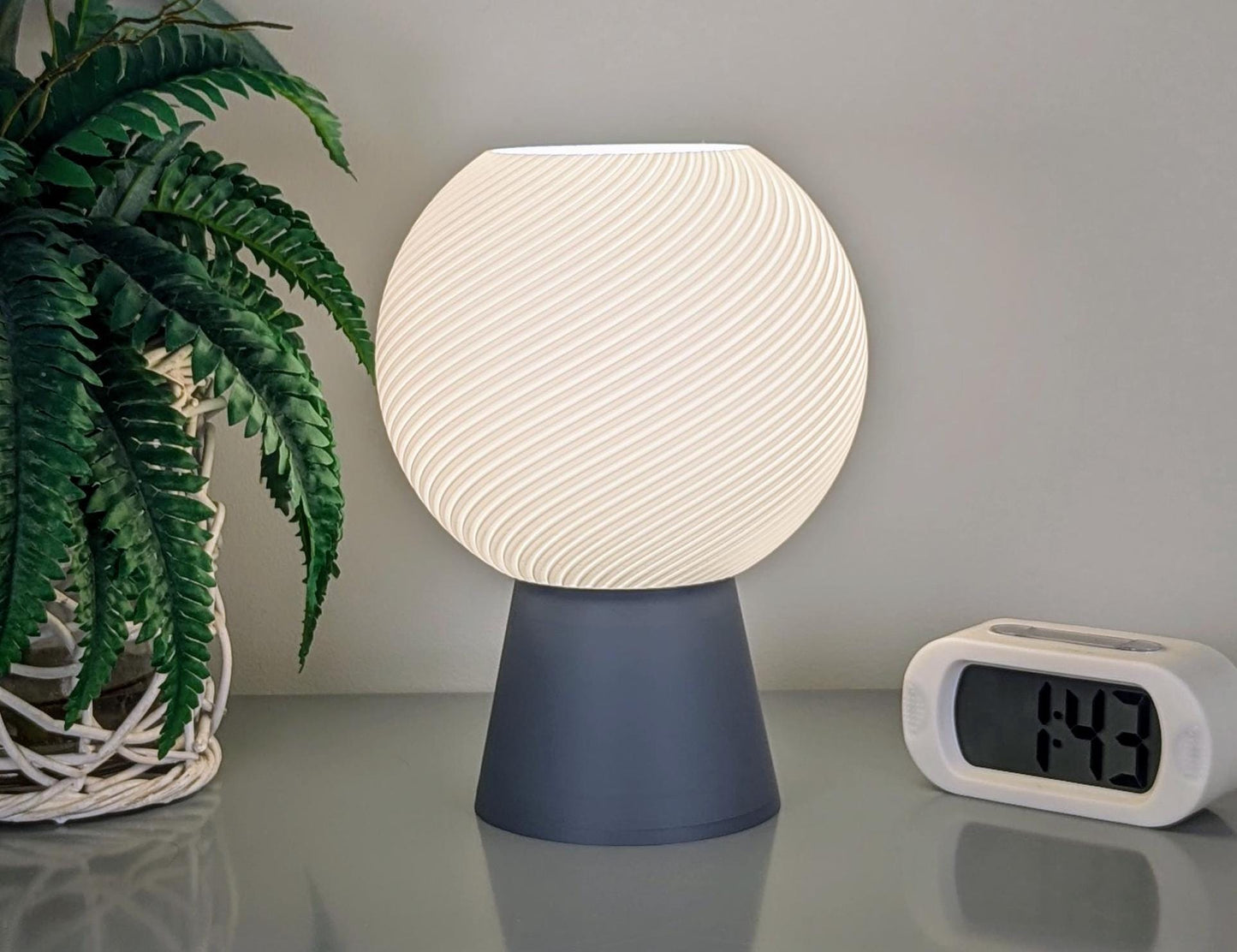 The Dryft Home Vortice Table Lamp, featuring a 3D-printed textured spherical shade, emits a soft glow next to a digital clock showing 1:43. Made from biodegradable PLA, it promotes sustainable lighting. A potted plant on the left adds natural charm to the setup.