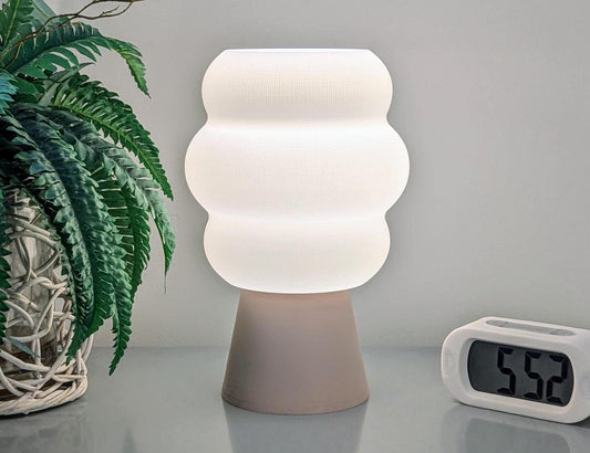 A Mammatus Table Lamp by Dryft Home, featuring a wavy white shade and sustainable craftsmanship, stands beside a digital clock showing 5:52. A green-leaved potted plant in a woven basket to the left completes the minimal, softly lit setting.