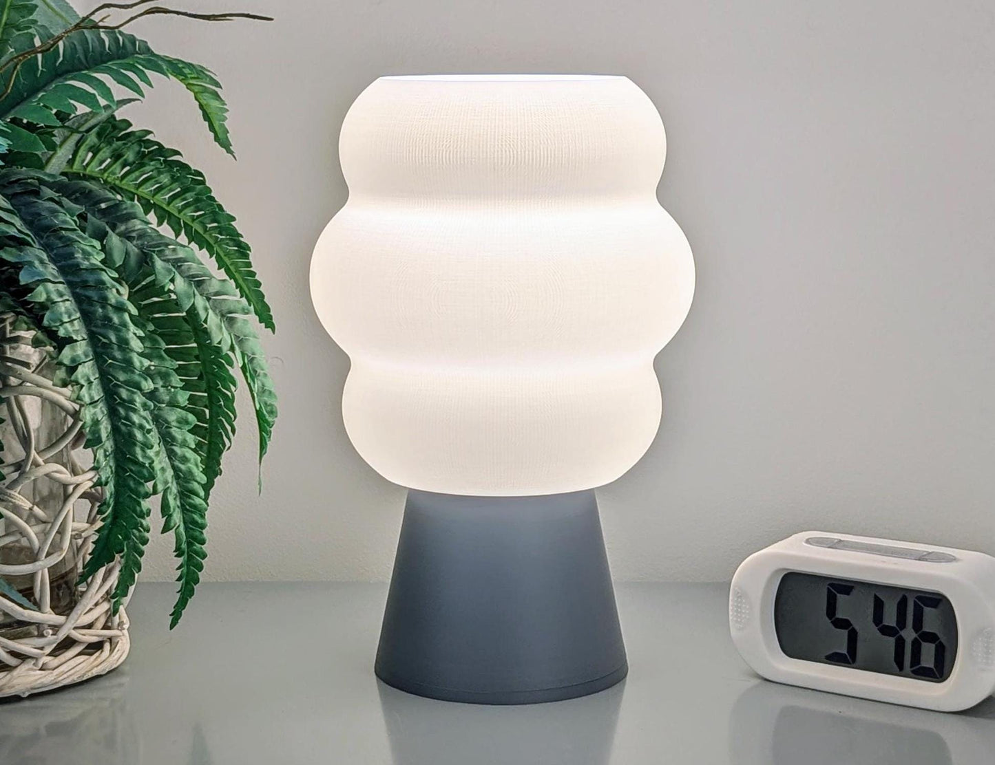 The Mammatus Table Lamp by Dryft Home, featuring a wavy white shade and crafted sustainably, is on a table beside a digital clock displaying 5:48. A woven basket holds a plant to the lamps left, enhancing the calm and minimalistic ambiance.