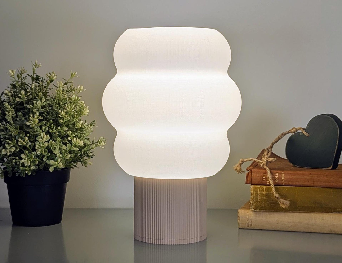 A Dryft Home Mammatus Table Lamp casts a warm glow on a table adorned with a small green plant, two stacked books, and a heart-shaped ornament tied with twine. This sustainable setup is displayed against a plain background.