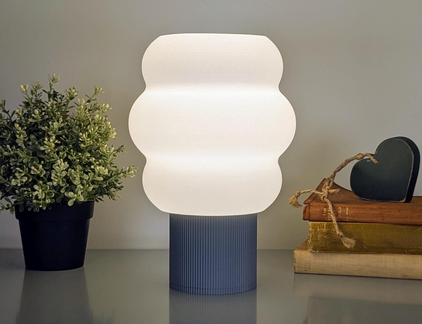 Dryft Homes Mammatus Table Lamp features a ribbed blue base and a wavy white shade, crafted sustainably. To its left is a potted plant with small green leaves, while on the right, books are topped with a green heart decoration.