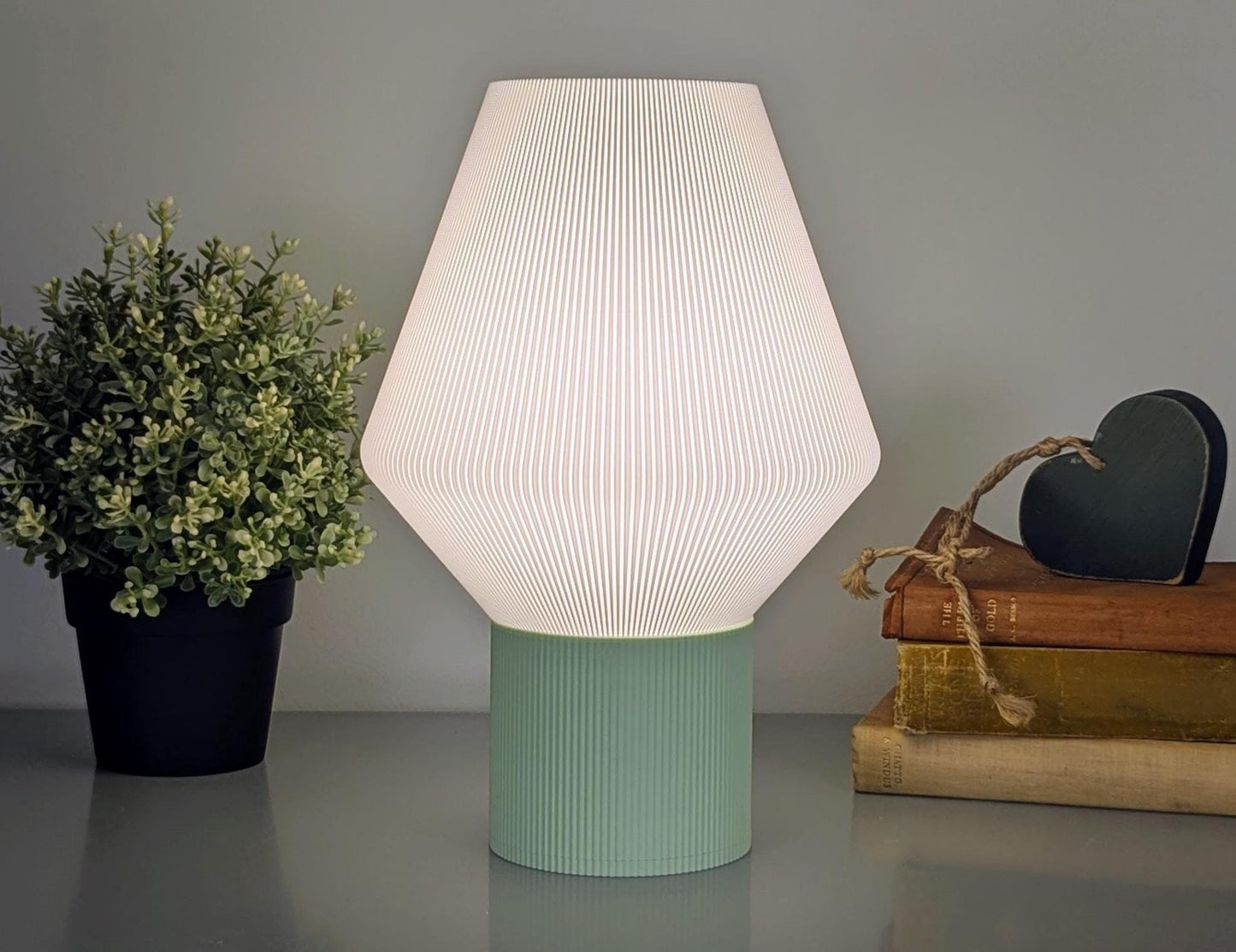 The Inversis Table Lamp by Dryft Home features a ribbed white hexagonal design, a light green base, and is made from eco-friendly PLA plastic. It sits on a surface next to a small potted plant and stacked books adorned with heart-shaped decor.