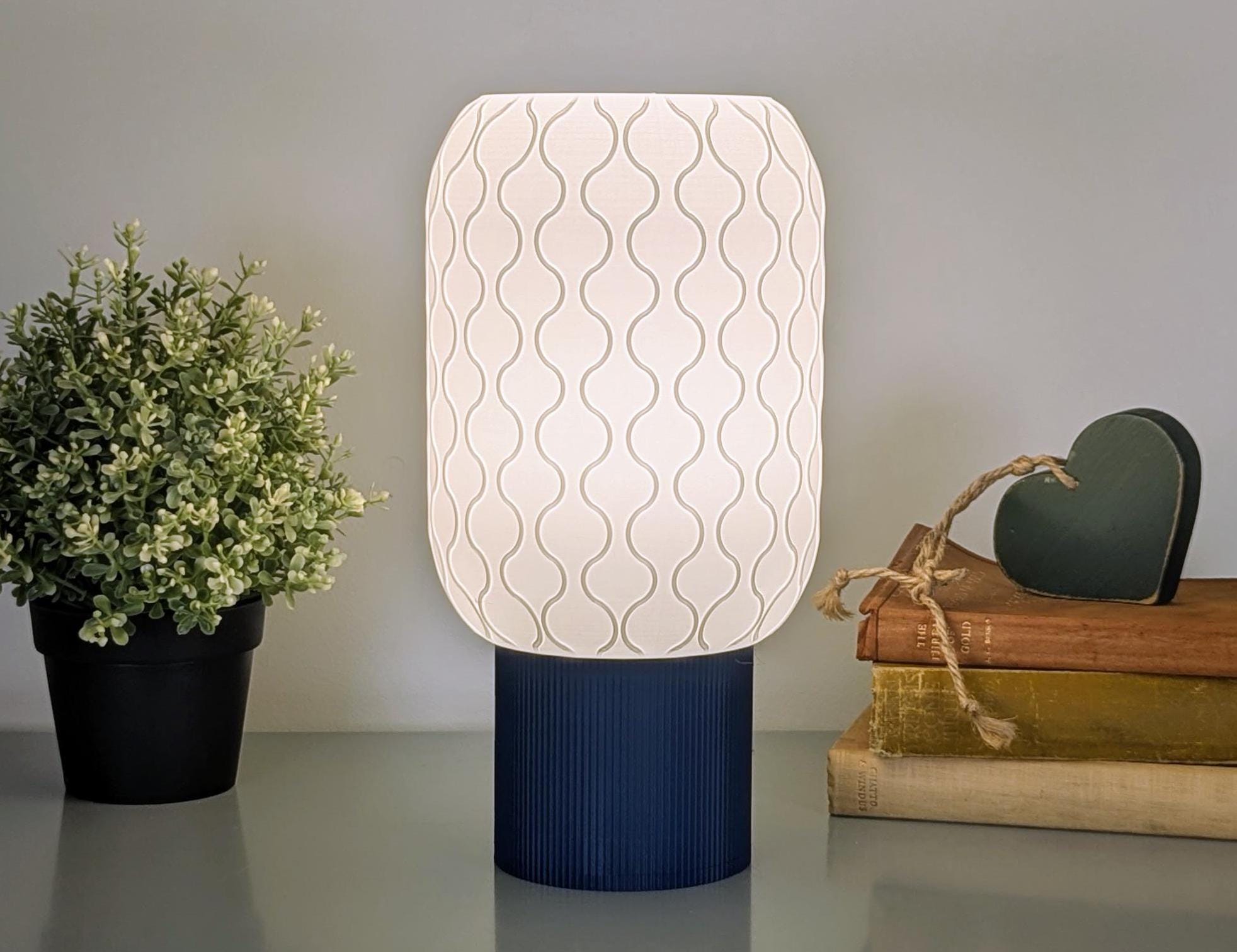 The Cirrus Table Lamp by Dryft Home, with its dark base and wavy shaded design, complements minimalist interiors. This 3D-printed piece offers sustainable beauty while sitting alongside a small potted plant and stacked books topped with a heart-shaped decoration on a surface.