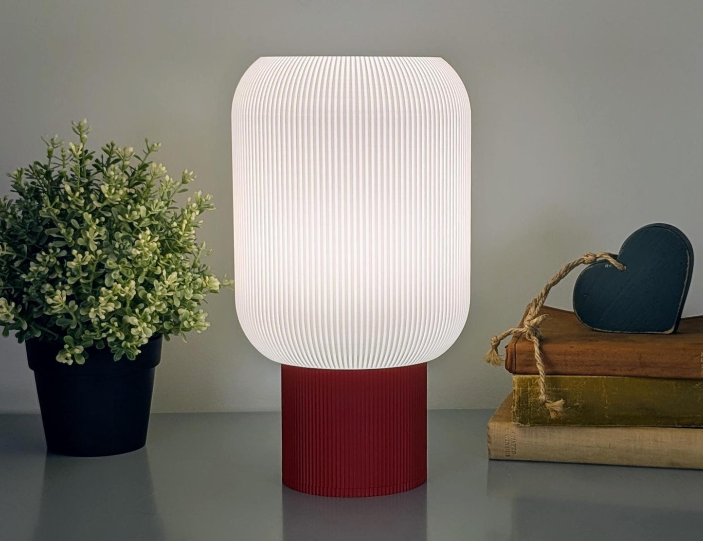 A warmly lit Cumulis Table Lamp by Dryft Home, crafted from sustainable materials with a 3D printed pleated cylinder, features a textured white shade and red base. To the left is a small potted plant, while to the right are stacked books with a wooden heart ornament.