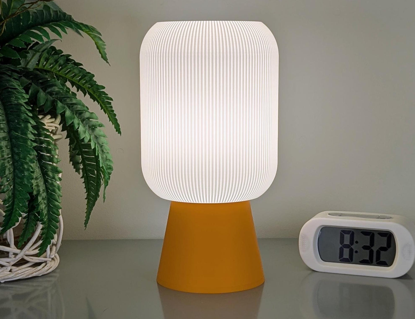 A Dryft Home Cumulis Table Lamp, featuring a 3D printed pleated white shade and vibrant yellow base made from sustainable materials, sits on a reflective surface. On the right is a clock showing 8:32, while a potted plant with feathery green leaves appears on the left.
