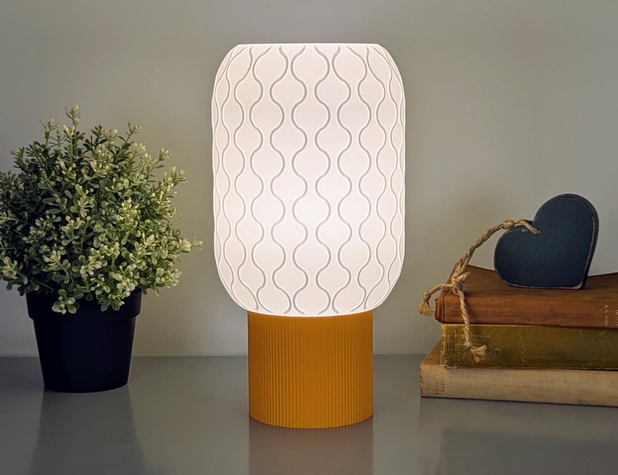 The Cirrus Table Lamp by Dryft Home features a modern 3D-printed design with an airy wavy shade and orange base, casting a warm glow. To its left sits a small potted plant, while to the right is a stack of books topped with a wooden heart decoration.