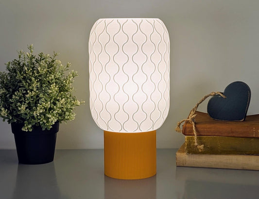 The Cirrus Table Lamp by Dryft Home features a modern 3D-printed design with an airy wavy shade and orange base, casting a warm glow. To its left sits a small potted plant, while to the right is a stack of books topped with a wooden heart decoration.
