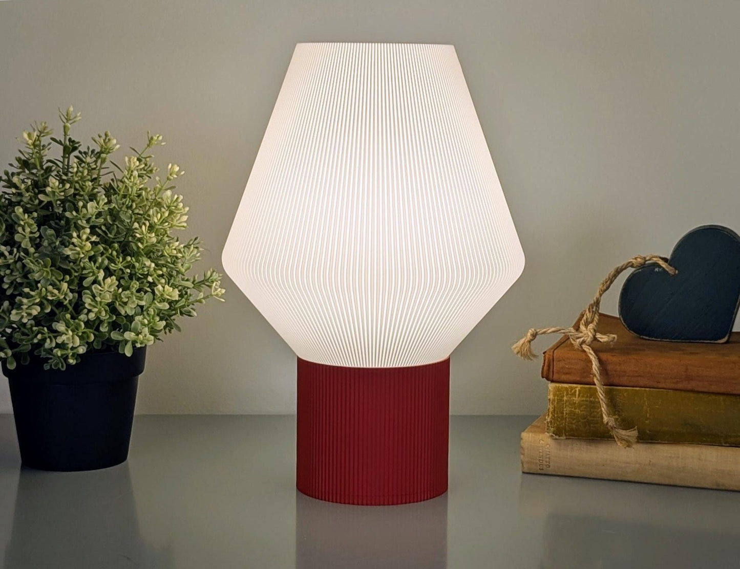 The Inversis Table Lamp by Dryft Home, a modern white lamp crafted from biodegradable PLA plastic and featuring a red base, sits on a surface. Beside it, a small plant and heart-decorated books emphasize sustainable production in your decor.