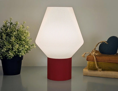 The Inversis Table Lamp by Dryft Home, a modern white lamp crafted from biodegradable PLA plastic and featuring a red base, sits on a surface. Beside it, a small plant and heart-decorated books emphasize sustainable production in your decor.