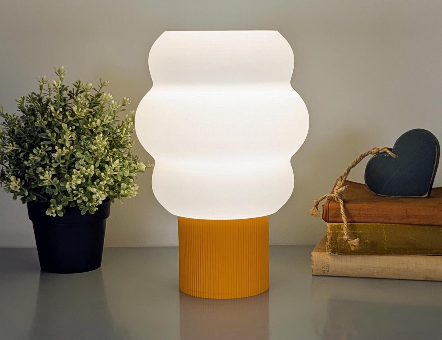 The Mammatus Table Lamp by Dryft Home, crafted using sustainable methods and biodegradable PLA plastic, features a white cloud-like shade on a yellow base. To the left is a green potted plant; to the right are stacked books with a dark heart-shaped ornament atop.