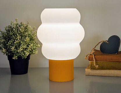 The Mammatus Table Lamp by Dryft Home, crafted using sustainable methods and biodegradable PLA plastic, features a white cloud-like shade on a yellow base. To the left is a green potted plant; to the right are stacked books with a dark heart-shaped ornament atop.
