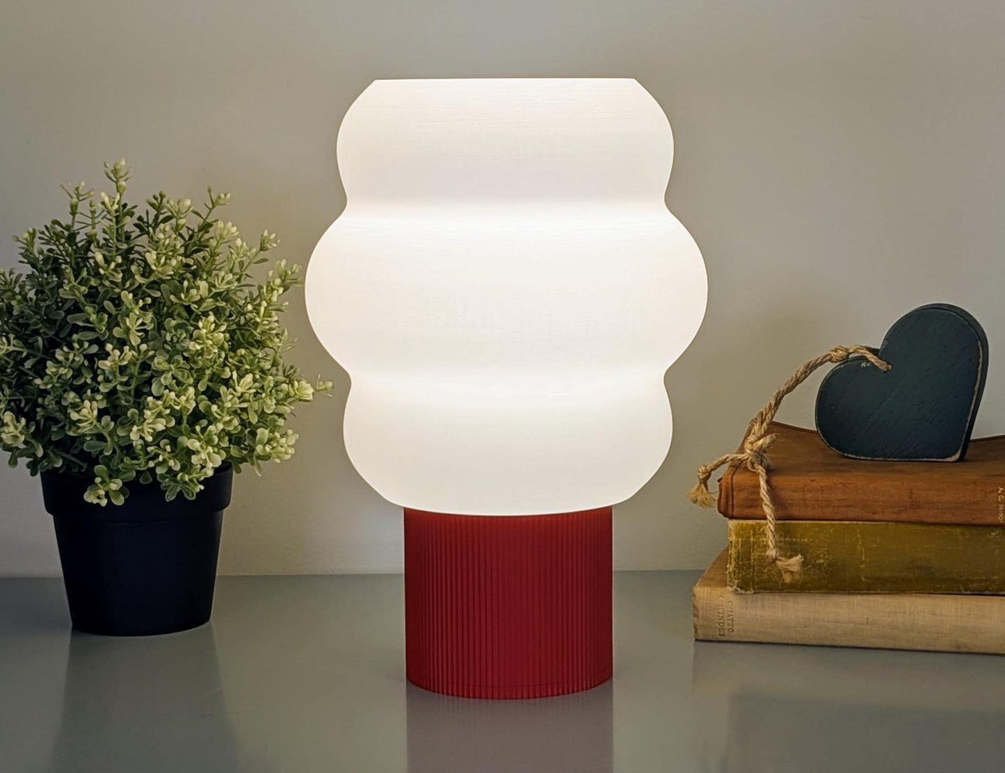 The Mammatus Table Lamp by Dryft Home, featuring a wavy white shade and vibrant red base, is eco-friendly with biodegradable PLA plastic. A small potted plant sits to its left, while two books with a heart ornament are on the right.