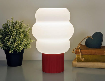 The Mammatus Table Lamp by Dryft Home, featuring a wavy white shade and vibrant red base, is eco-friendly with biodegradable PLA plastic. A small potted plant sits to its left, while two books with a heart ornament are on the right.