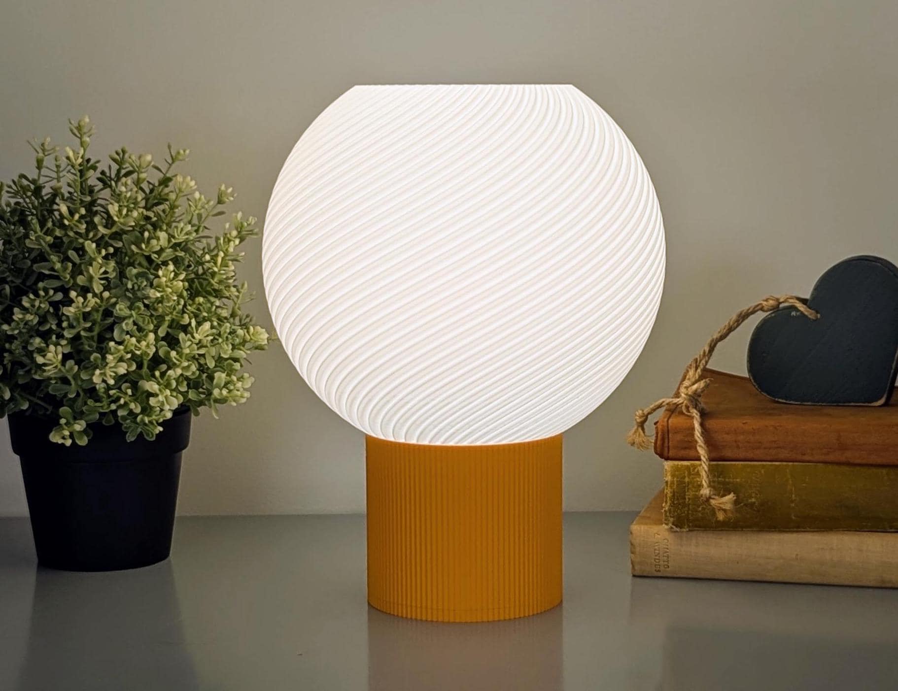 The Vortice Table Lamp by Dryft Home, with a textured white biodegradable PLA plastic shade and orange base, glows on a table. Nearby, a small potted plant sits next to books topped with a heart-shaped decor piece.