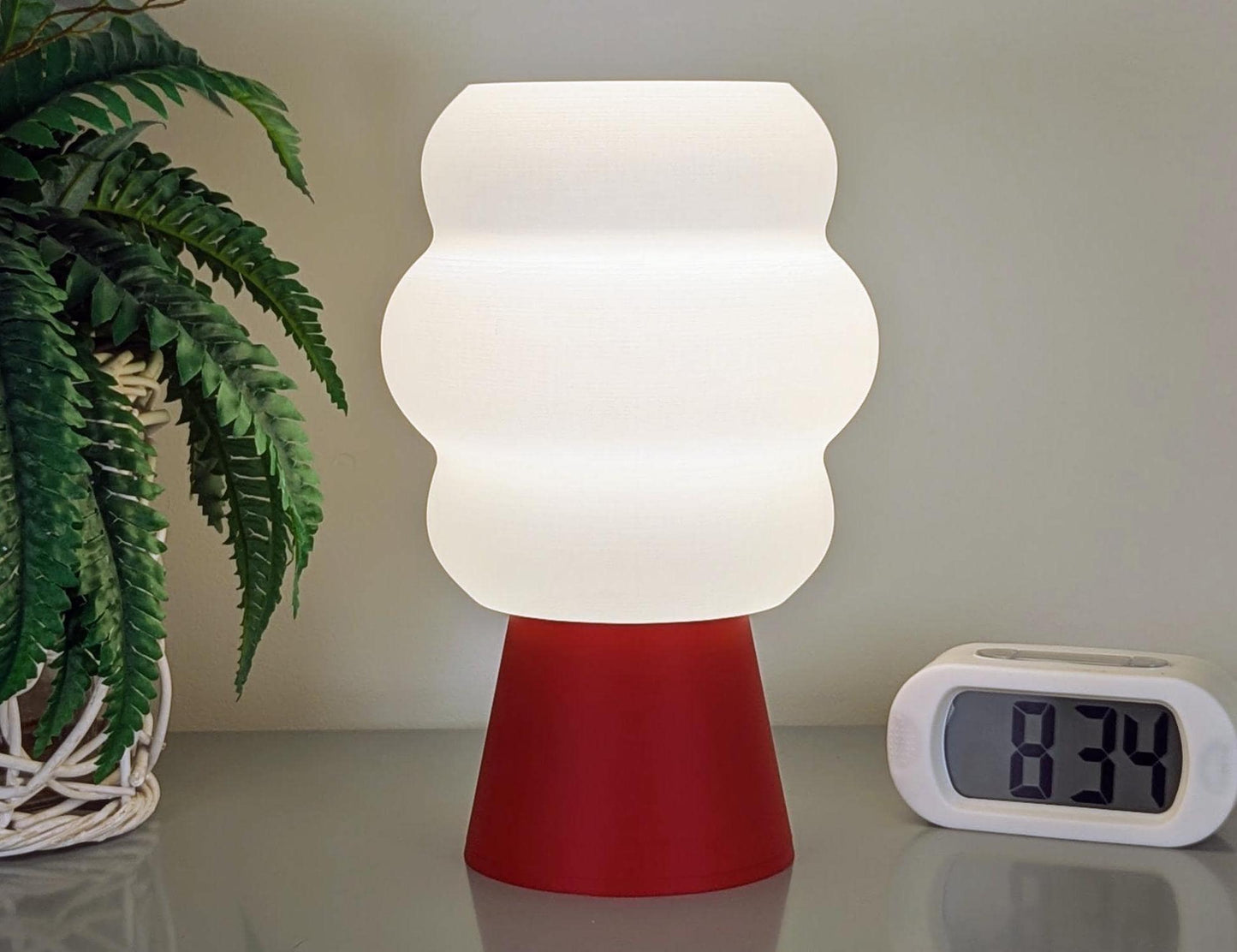 The Mammatus Table Lamp by Dryft Home, 3D printed from PLA plastic, elegantly rests on a surface with its red base. It casts a glow next to a digital clock showing 8:34, while the green fronds of a potted plant add natural charm to this sustainable scene.