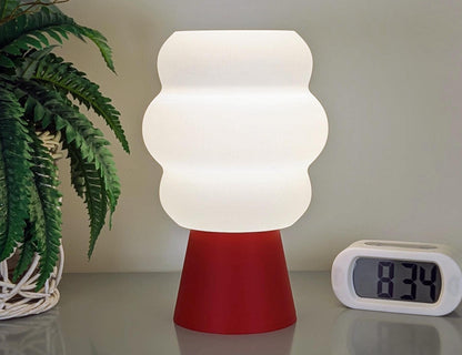 The Mammatus Table Lamp by Dryft Home, 3D printed from PLA plastic, elegantly rests on a surface with its red base. It casts a glow next to a digital clock showing 8:34, while the green fronds of a potted plant add natural charm to this sustainable scene.