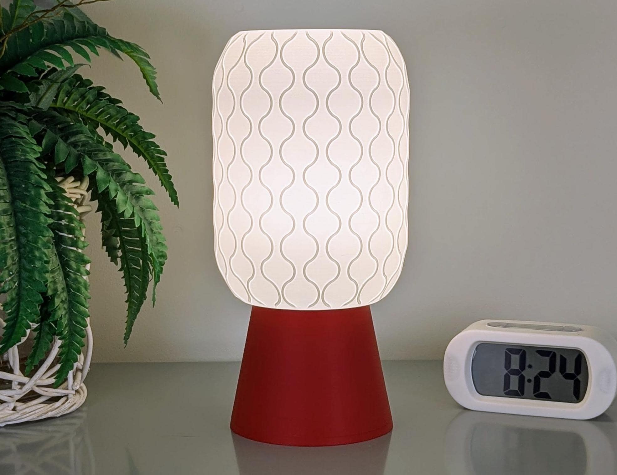 The Cirrus Table Lamp – 3D Printed Airy Elegance by Dryft Home, featuring a wavy shade and red base, sits on a table beside an 8:24 digital clock. In the background, a leafy plant in a wicker basket enhances the minimalist interior style.