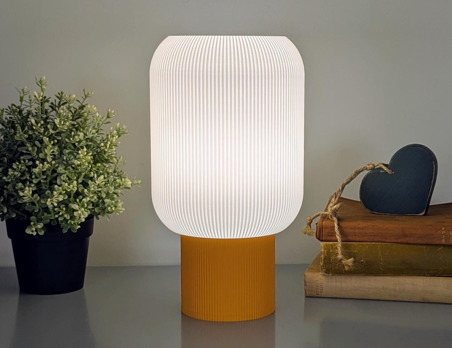 The Cumulis Table Lamp – 3D Printed Pleated Cylinder Lighting by Dryft Home, made from sustainable materials, features a ribbed white shade and mustard base. It softly illuminates over stacked books with a heart-shaped ornament on the right and a small potted plant on the left.