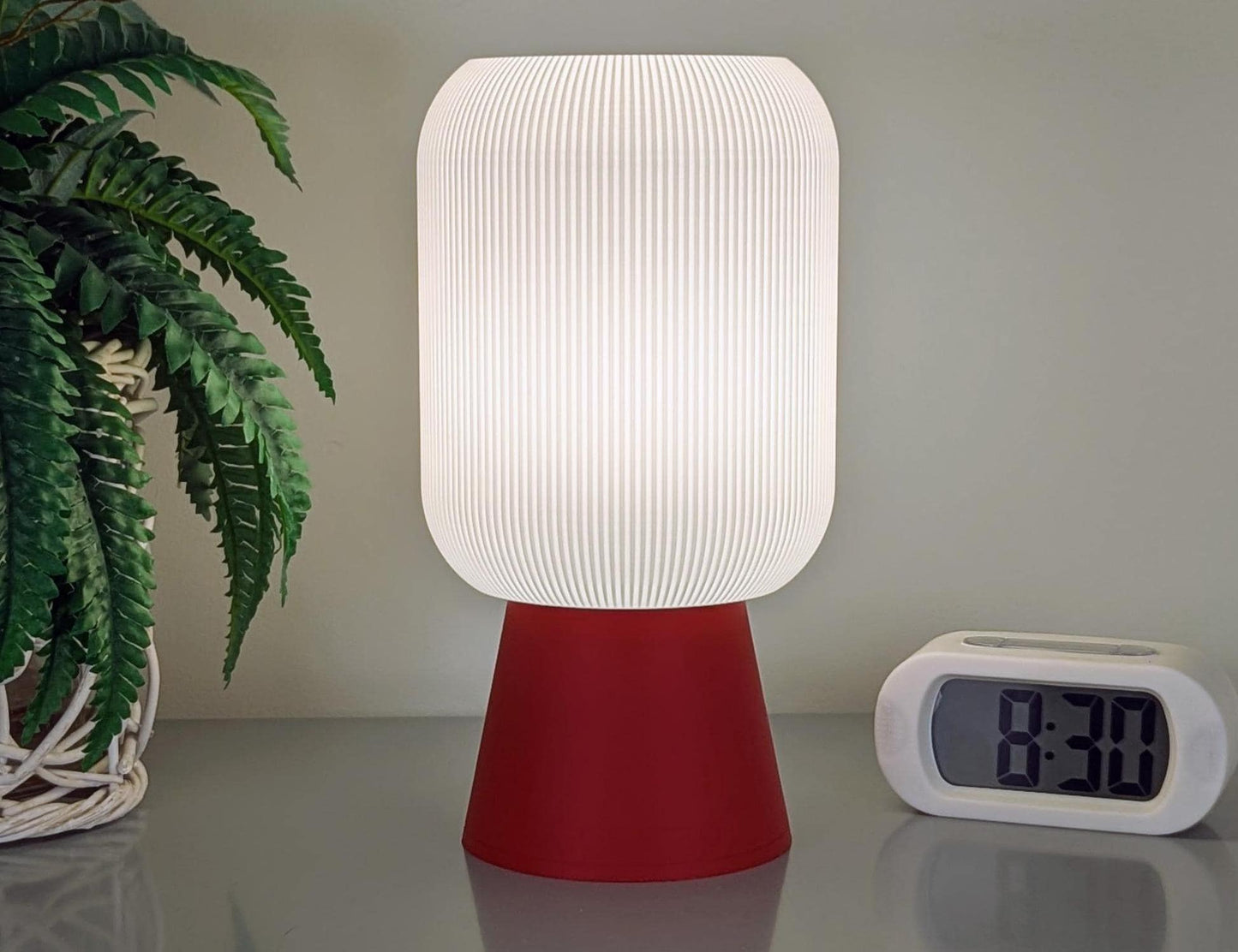 A Dryft Home Cumulis Table Lamp, in 3D printed pleated cylinder design with a red base and eco-friendly materials, brightens a gray surface. To its right, a digital clock displays 8:30 as a potted plant with green leaves peeks in from the left.