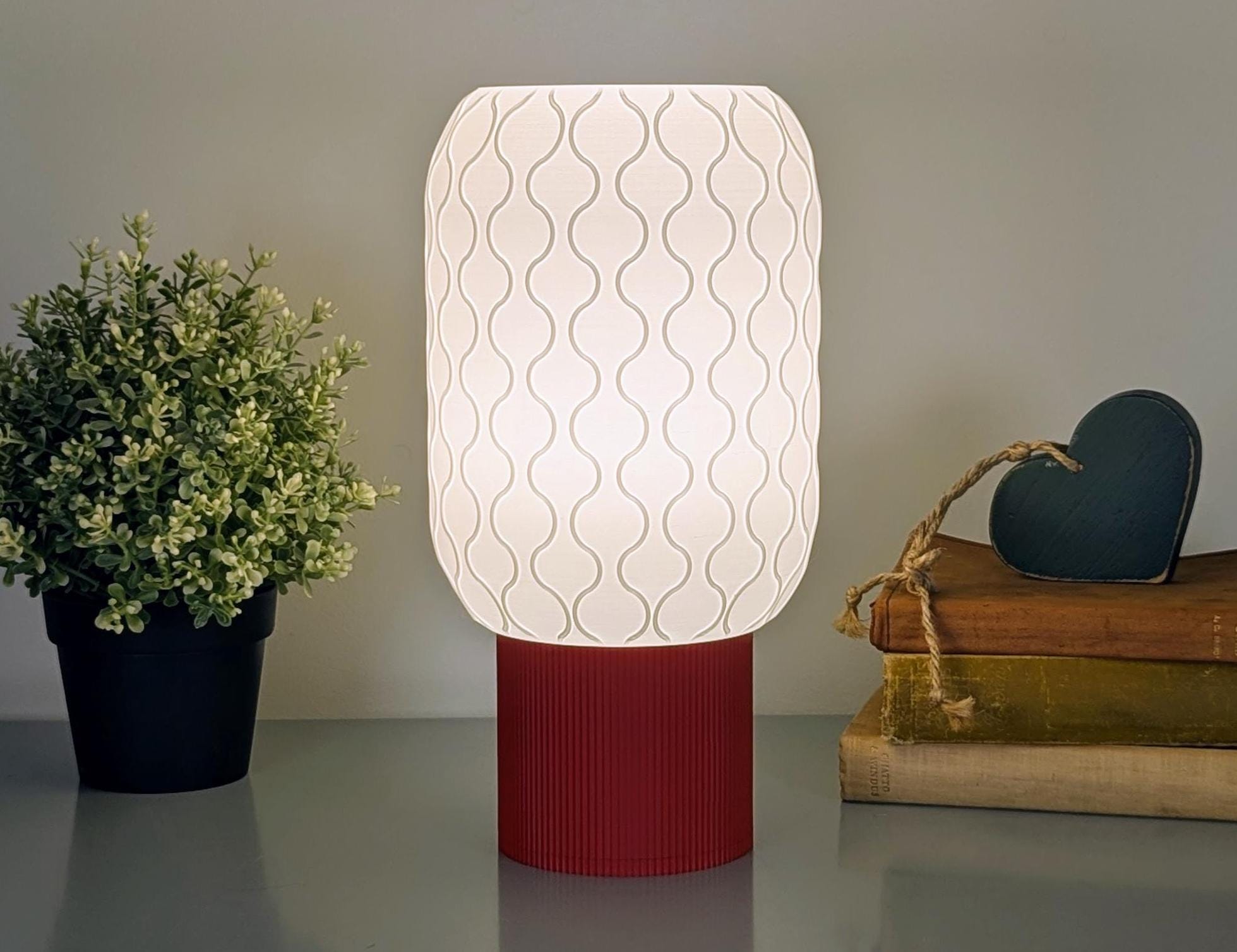 The Cirrus Table Lamp by Dryft Home, featuring a wavy-patterned shade and red base, is perfect for minimalist interiors. It sits among a small potted plant to the left and stacked books with a dark heart-shaped ornament to the right, showcasing sustainable materials.