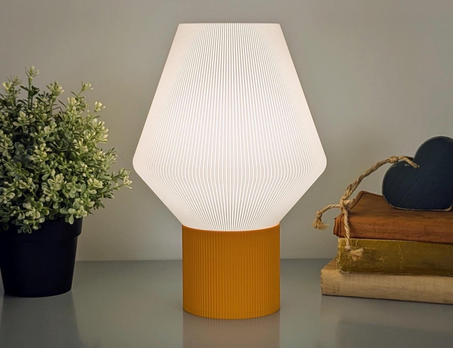 The Inversis Table Lamp by Dryft Home is a modern, geometric piece 3D printed from biodegradable PLA plastic. It features a bright white conical shade and an orange base, complemented by a lush potted plant and stacked books with a heart-shaped decorative object, highlighting sustainable production choices.