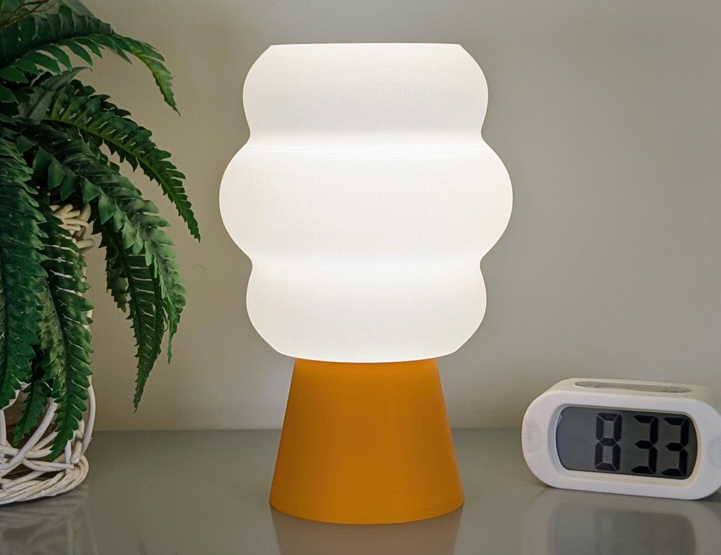 The Dryft Home Mammatus Table Lamp, 3D printed with a wavy white shade from eco-friendly PLA and an orange base, sits beside a digital alarm clock displaying 8:33. A green potted plant in the background highlights the fusion of technology and sustainable design.