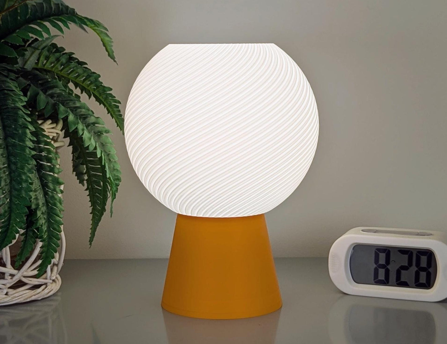The Vortice Table Lamp from Dryft Home, crafted with biodegradable PLA, showcases a white textured spherical shade and a yellow base elegantly sitting on a table. A potted plant with green fronds is to its left, and a digital clock displays 8:28 on its right.