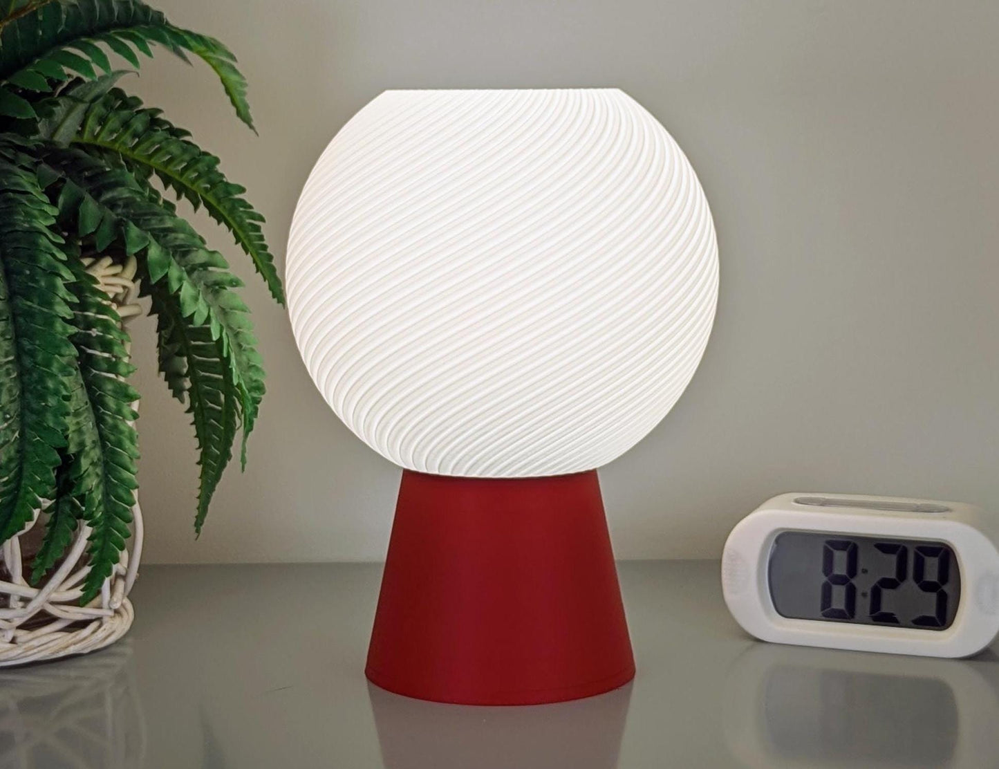 The Vortice Table Lamp by Dryft Home, made from biodegradable PLA with a spiral texture, has a glowing white shade on a red base. Its elegantly set next to a digital clock displaying 8:29 and an indoor plant with lush green leaves.
