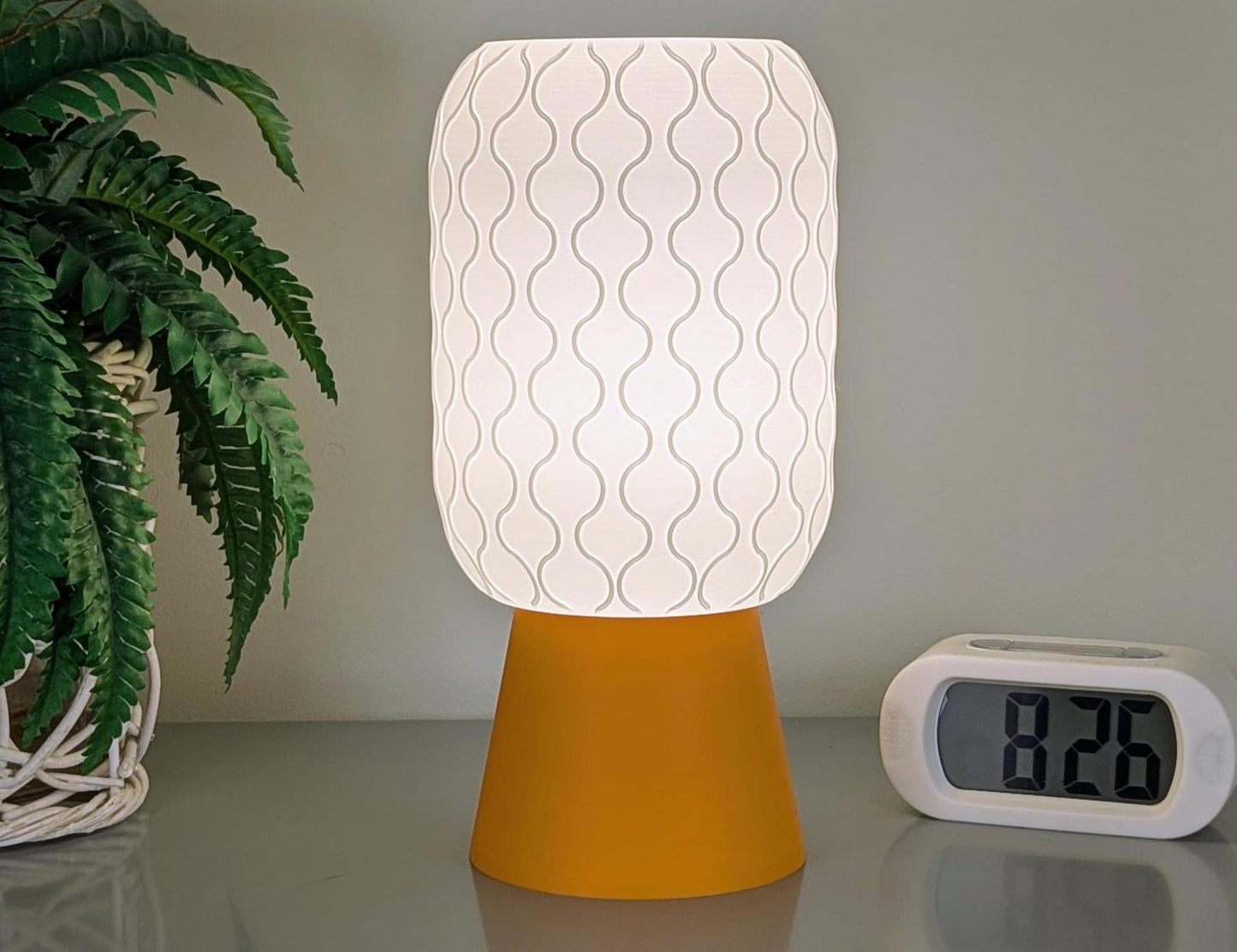 The Cirrus Table Lamp by Dryft Home, with its 3D-printed wavy white shade and yellow base, graces the surface. At 8:20, a digital clock is seen to the right. Minimalist interiors get a touch of sustainable elegance as green fern-like foliage appears on the left.